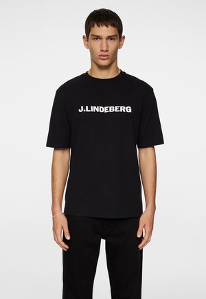 Mustan puuvillan T-paita lyhyillä hihoilla, jossa on valkoinen, rohkea "J.LINDEBERG" -logo rinnan kohdalla. Yksinkertainen muotoilu, normaali istuvuus.
