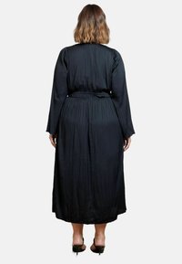 Svart plisserad klänning med långa ärmar och bälte i midjan, med en flödande silhuett och mjuk textur, som når till mitten av vaderna.