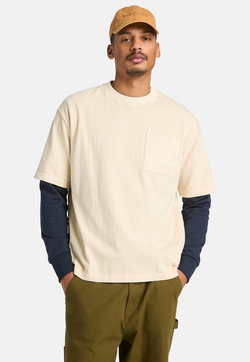 Homme portant une chemise beige à manches courtes par-dessus une chemise bleu marine à manches longues, un pantalon olive et une casquette Timberland beige, debout les mains dans les poches.