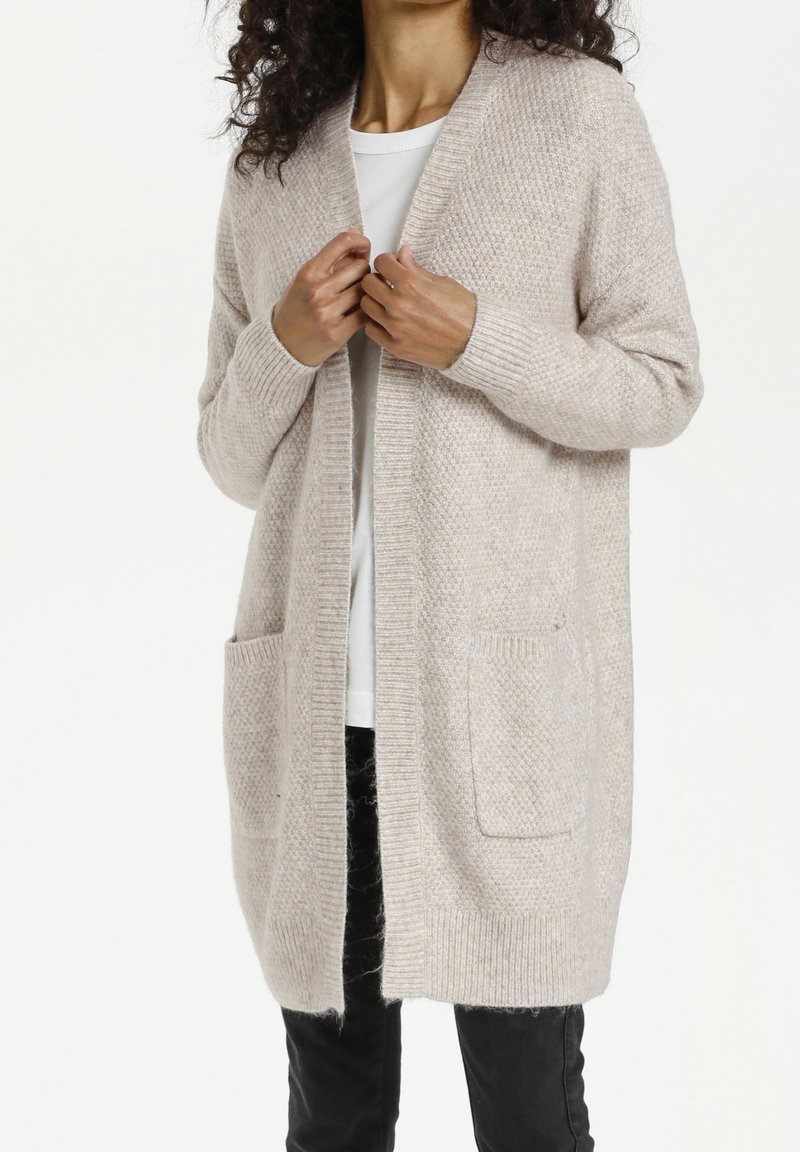 Femme portant un long cardigan beige en maille avec des poches frontales par-dessus une chemise blanche et un pantalon noir, les mains ajustant le col du cardigan.