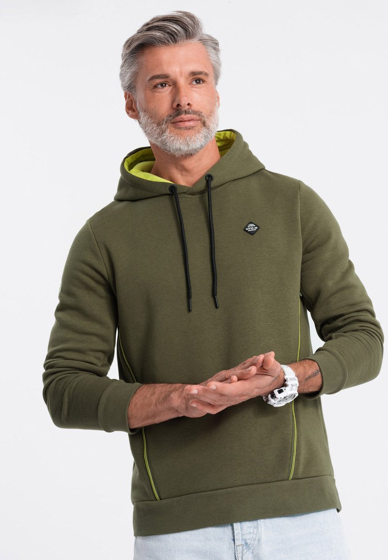 Sudadera verde oliva con un logo negro, cordones ajustables y forro amarillo brillante. Cuenta con detalles laterales y puños acanalados.