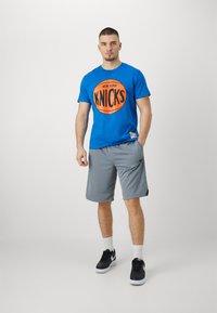 Jasnoniebieska bawełniana koszulka z okrągłą pomarańczową grafiką "NEW YORK KNICKS", w zestawie z szarymi szortami sportowymi i czarnymi sneakersami.
