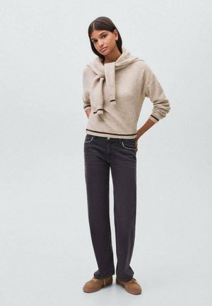 Beige sweater med ribbet kant og hals, sikret med en knude; parret med mørke jeans med sølvdetaljer på lommerne og beige sko.