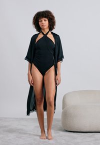 Melna satīna halāta ar mežģīņu apdari, kas valkāts virs melna viengabala peldkostīma. Halāts ir ar atvērtu priekšu un plūstošām piedurknēm, savienots ar kailām kājām.