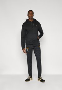 Μαύρο φούτερ με κουκούλα και μπροστινή τσέπη, συνδυασμένο με μαύρο παντελόνι με σχέδιο και λογότυπο UFC. Μαύρα παπούτσια με χρυσές λεπτομέρειες.