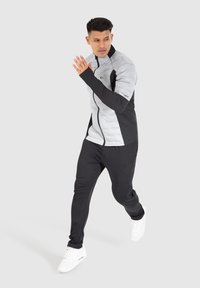 Grå och svart träningsjacka med dragkedja och långa ärmar, ihopparad med svarta jogger-byxor och vita sneakers, mot en neutral bakgrund.