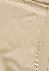 Bézs színű, sima textúrájú, bordázott mintázatú szövetleggings ezüstös fényű logóval az oldalán.