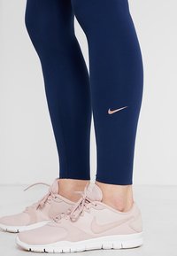 Námořnické legíny z pružného materiálu s přiléhavým střihem, s malým růžovým zlatým logem Nike na dolní části nohy, v kombinaci se světle růžovými teniskami.