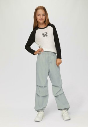 Junges Mädchen steht mit einer Hand in der Hüfte, trägt ein schwarzes und weißes Langarmshirt mit Schmetterlingsdruck, lockere hellgraue Hosen und weiße Turnschuhe.