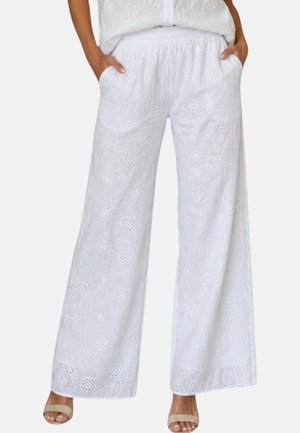Pantaloni bianchi a gamba larga con motivo floreale traforato, vita elasticizzata e tasche, indossati con sandali beige aperti e mani nelle tasche.