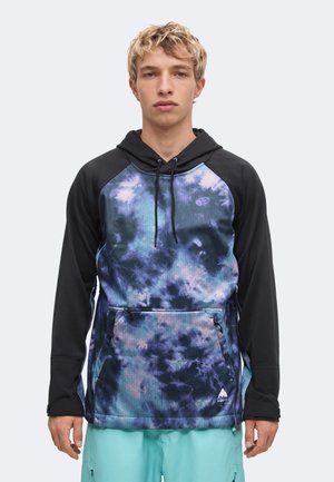 Joven con una sudadera con capucha de mangas negras, parte delantera en azul y púrpura con efecto tie-dye y pantalones turquesa, de pie frente a un fondo liso y claro.