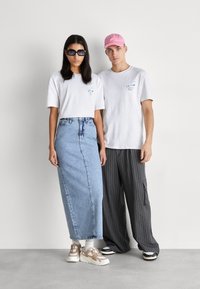 Dos modelos están uno al lado del otro. La mujer lleva una camiseta corta blanca, una falda de mezclilla y gafas de sol. El hombre lleva una camiseta blanca, pantalones a rayas y una gorra rosa.