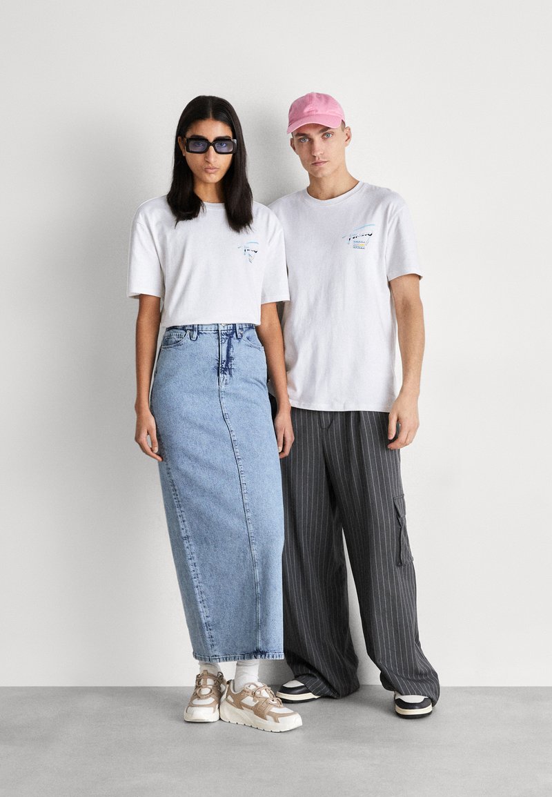 Dos modelos están uno al lado del otro. La mujer lleva una camiseta corta blanca, una falda de mezclilla y gafas de sol. El hombre lleva una camiseta blanca, pantalones a rayas y una gorra rosa.