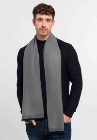 Écharpe en chevrons grise en matériau doux, avec des extrémités effilochées et un éclat subtil, portée enroulée autour du cou.