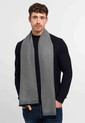 Gray herringbone scarf made of soft material, featuring fringed ends and a subtle sheen, worn draped around the neck.