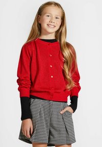 Czerwony kardigan z błyszczącymi akcentami, okrągłym dekoltem i zapięciem na guziki. Czarne ściągacze rękawów. Połączony z szortami w czarno-białą kratę houndstooth.