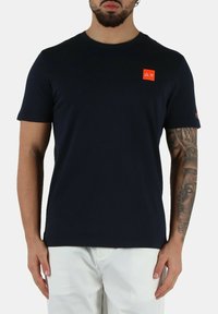 T-shirt a maniche corte blu navy in un tessuto liscio, con una piccola toppa con logo arancione sul petto e un accento rosso sulla manica.