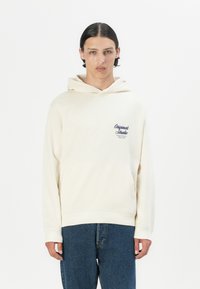 Hoodie color crema con tasca frontale e logo ricamato sul lato sinistro; tessuto texturizzato; abbinato a jeans blu, design minimalista.
