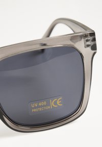 Lunettes de soleil noires et grises avec une épaisse monture rectangulaire. Verres foncés étiquetés "PROTECTION UV 400 ICE". Texture lisse et accents clairs.