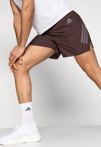 Καφέ αθλητικό σορτς με γκρι σχεδιασμό τριών ρίγων, ελαφρύ υλικό, πλαϊνές σχισμές, σε συνδυασμό με λευκές κάλτσες και παπούτσια.