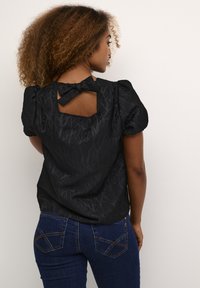 Zwarte textuurblouse met pofmouwen en een uitsnede aan de achterkant met een strikdetail. Draag het met donkerblauwe jeans met decoratieve stiksels.