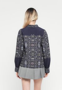 Woman with long blonde hair wears dark floral long-sleeve blouse and light gray pleated skirt, standing against a white background.