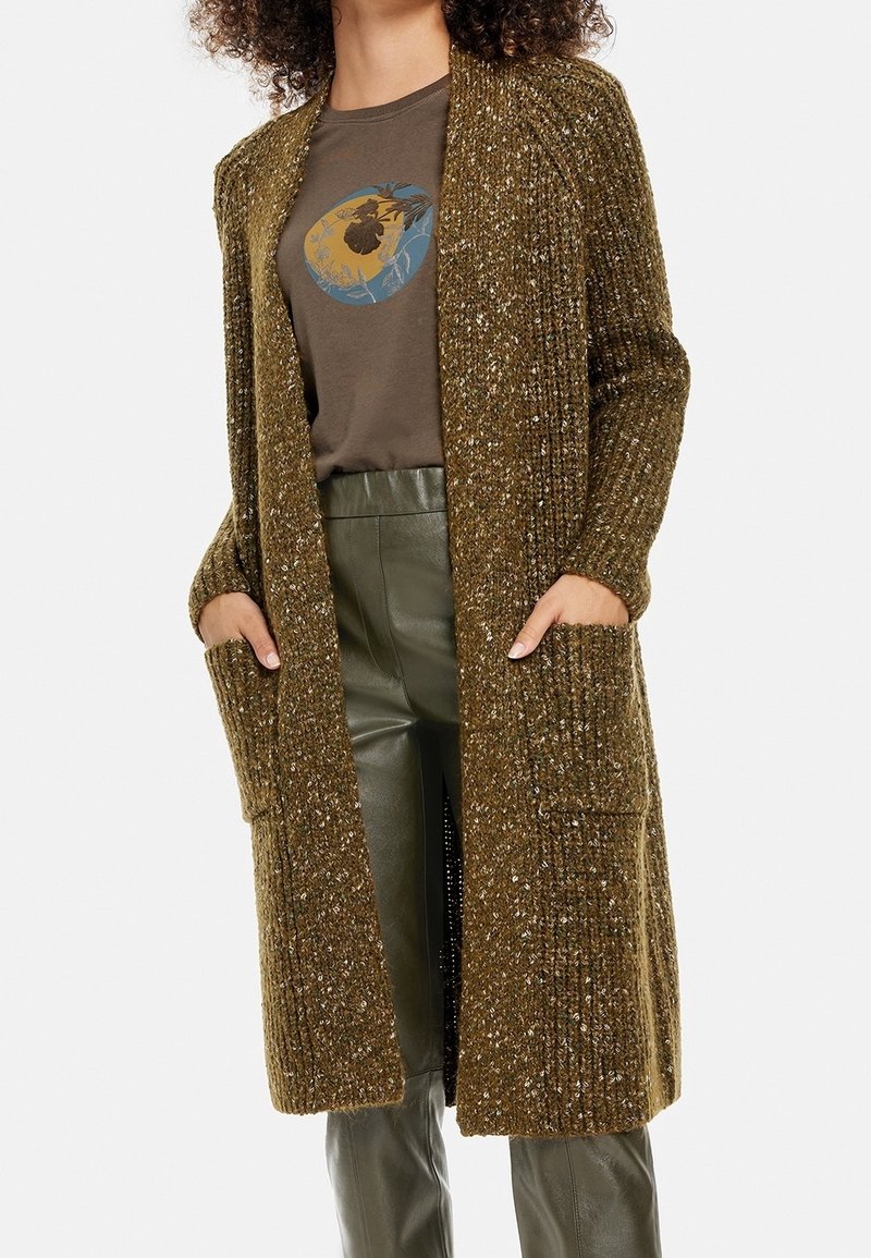 Strickjacke in Olivgrün mit einem strukturierten Muster, langen Ärmeln, offenem Frontbereich und zwei vorderen Taschen, getragen über einem grafischen T-Shirt.