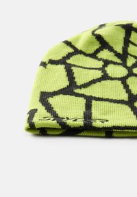 Bonnet en tricot vert avec un motif de toile d'araignée géométrique noir. Le matériau est un tissu côtelé texturé avec un détail d'ourlet replié.