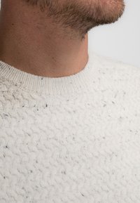 Textured white sweater with a wavy knit pattern, featuring a ribbed crew neck and subtle darker flecks throughout the fabric.