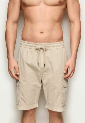 Mężczyzna ubrany w beżowe szorty bojówki z sznurkiem w pasie, stojący z opuszczonymi rękami na tle neutralnym.