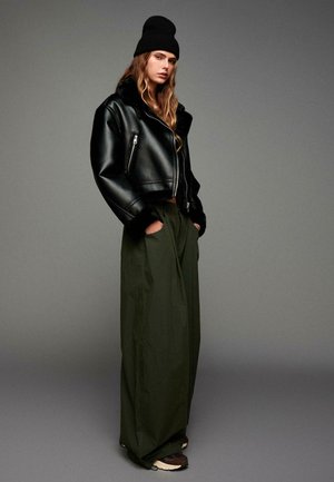 Black leather cropped jacket with faux fur collar, paired with olive green wide-leg pants and dark brown sneakers. Black beanie on head.