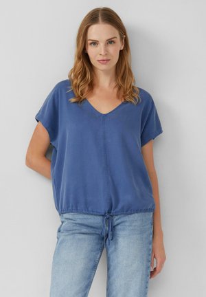 Blusa blu a maniche corte con scollatura a V, orlo arricciato e dettaglio con coulisse. Tessuto morbido con una consistenza liscia. Abbinata a jeans chiari.