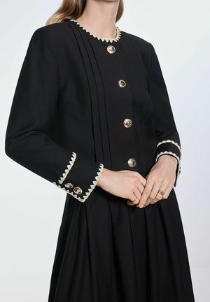 Vrouw draagt een zwarte jurk met witte zigzagbies op de halslijn en manchetten, en gouden knopen aan de voorkant en op de mouwen.