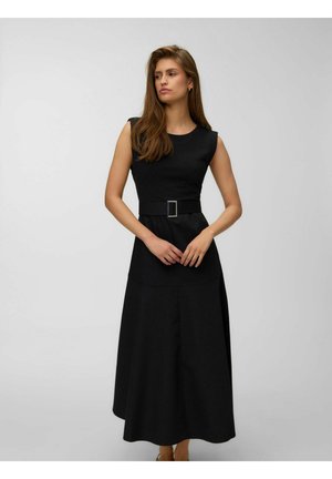 Femme en robe noire sans manches avec ceinture, debout devant un fond gris clair uni, regardant sur le côté les mains croisées.