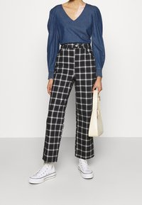 Top in denim con scollatura a V e maniche a palloncino, abbinato a pantaloni a quadri bianchi e neri con vita alta e sneakers bianche. Tiene una borsa color crema.