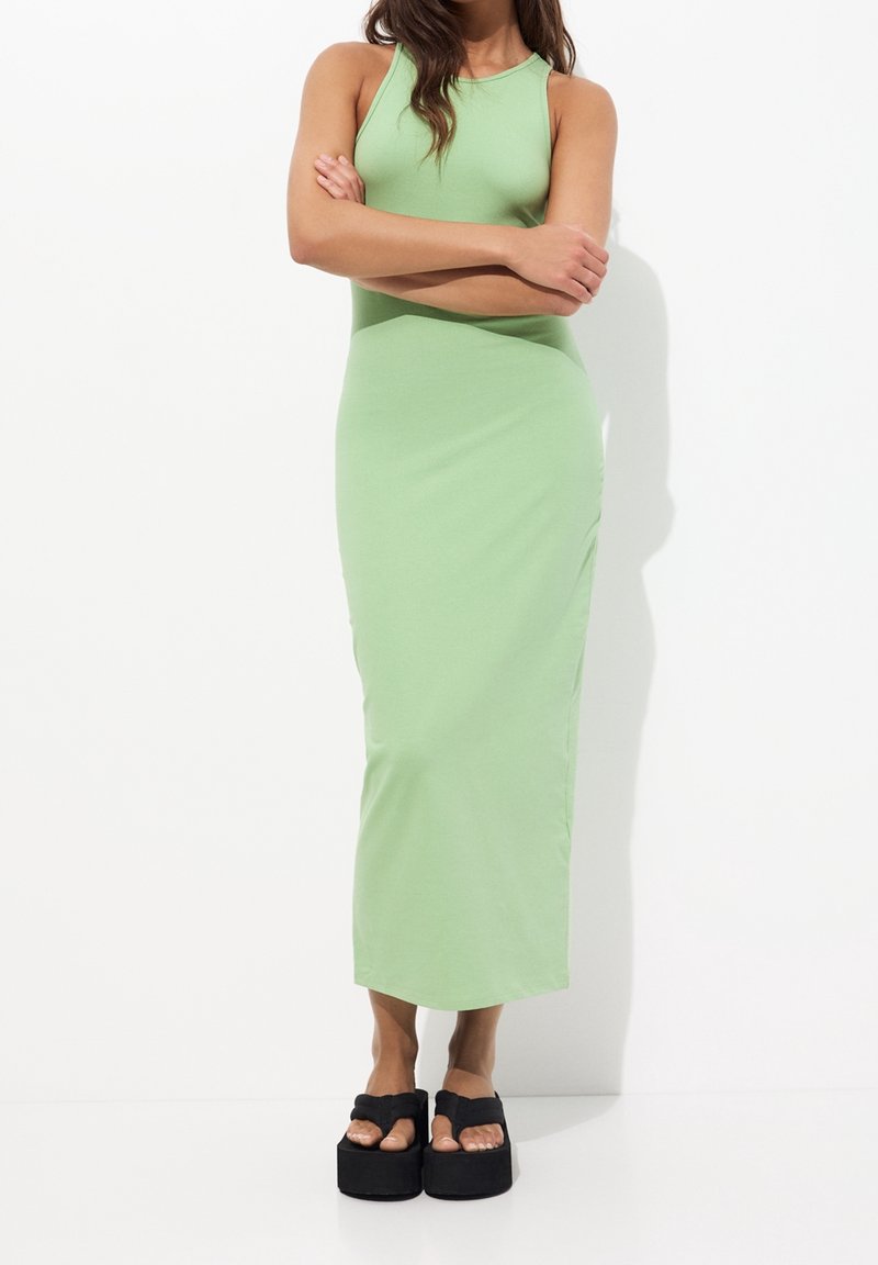 Kobieta stoi z założonymi na sobie rękami, ubrana w bezrękawową, jas zieloną maxi sukienkę oraz czarne platformy, na tle białego tła.