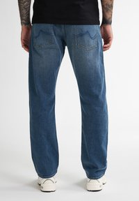 Blauwe spijkerbroeken met een rechte pijp, vervaagde was en twee achterzakken. Draagt men met witte sportschoenen. Minimale details.