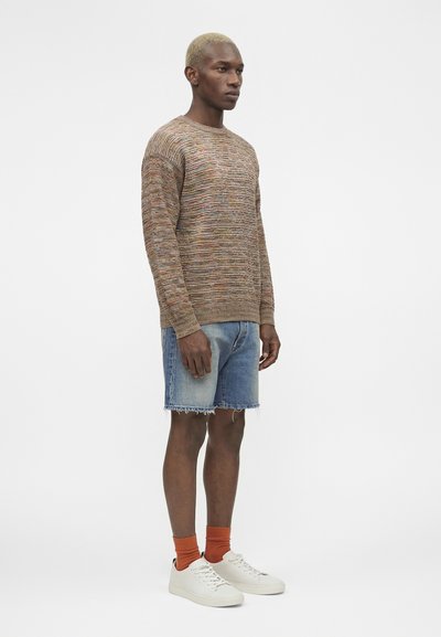 Άνδρας με κοντά ξανθά μαλλιά που φοράει πολύχρωμο πλεκτό πουλόβερ, ξεθωριασμένα τζιν σορτς, πορτοκαλί κάλτσες και λευκά αθλητικά παπούτσια, στέκεται πλαγίως.