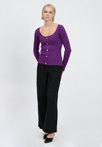 Lila gerippte Langarmbluse mit einem runden Ausschnitt, ausgestattet mit goldenen quadratischen Knöpfen vorne. Kombiniert mit schwarzen weit geschnittenen Hosen.
