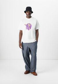 T-shirt en coton blanc avec un design graphique violet, associé à un pantalon en denim gris ample et des chaussures en cuir marron, le tout complété par un chapeau seau noir.