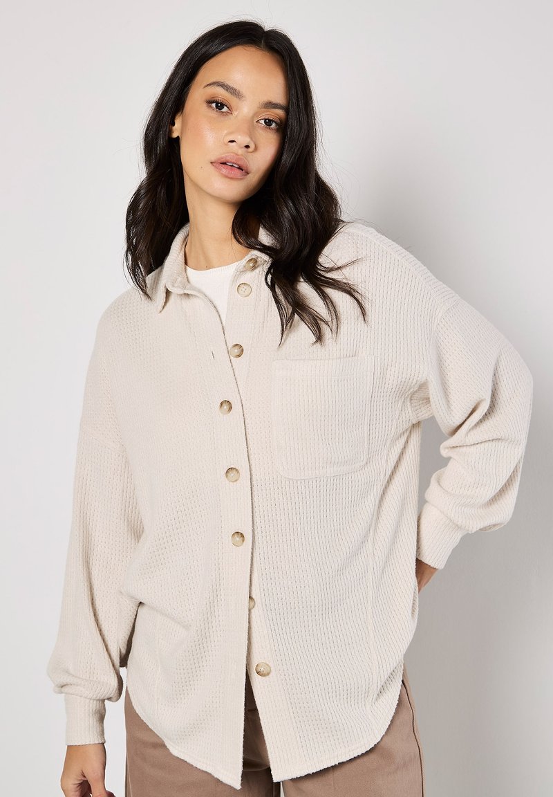 Waffle-knit beige button-up shirt with a collared neckline, long sleeves, chest pocket, and rounded hem. Light brown buttons contrast the fabric.
