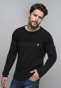 Maglione nero lavorato a maglia con un motivo testurizzato, dotato di un giromanica a costine e maniche lunghe. Piccolo logo decorativo sul petto. Indossato con jeans.