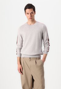 Pull en maille gris clair avec des poignets et un ourlet côtelés, présentant des accents rayés bleu marine et rouge sur les manches. Assorti à un pantalon beige.