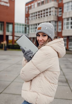 Woman in beige puffer jacket, gray knit hat, and gloves holding a light gray wallet outdoors with modern building in the background.