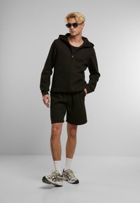 Sudadera negra con cremallera y capucha, acompañada de pantalones cortos negros. El modelo lleva gafas de sol y zapatillas deportivas con calcetines blancos.