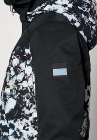 Manche de veste noire avec un patch rectangulaire bleu du logo Roxy, tissu à motif floral sur le corps et une partie de la capuche visible.
