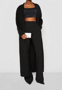 Un long cardigan noir, un top noir court, un pantalon noir taille haute et des talons aiguilles noirs. Tient un portefeuille CK blanc avec un motif matelassé.