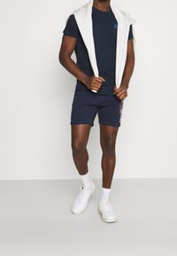 Navy t-shirt med logotyp, matchat med navy shorts med sidobranning. Vit tröja över axlarna, vita sneakers och ankelsockor.
