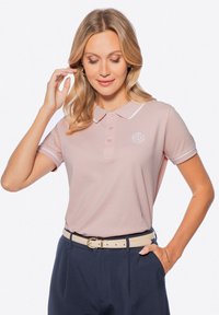 Světle růžová polo košile s bílým lemem a logem, s límečkem a krátkými rukávy. Nošena s námořnicky modrými kalhotami a béžovým páskem.
