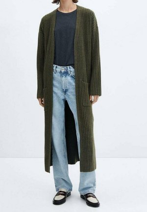 Osoba oblečená v dlouhém olivově zeleném žebrovaném svetru, tmavě šedém topu, světle modrých džínách a černo-bílých mokasínách, stojící před bílým pozadím.